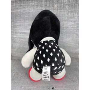 May include: A soft toy with a black head and a body patterned with black and white diamonds. It has white arms and red feet. A tag with the word "LAIKA" is attached to the toy.