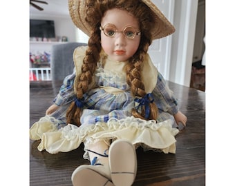 Puede incluir: Una muñeca de porcelana con el pelo castaño en trenzas, que lleva un vestido de cuadros azules y blancos con ribete de encaje amarillo, un sombrero de paja y gafas redondas.