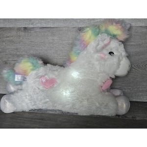 May include: A white plush unicorn toy with a rainbow mane and tail. The unicorn has a pink heart-shaped patch on its side and glowing lights. The hooves are silver and glittery. A tag is visible on the side.