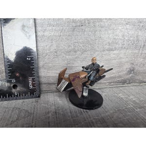 May include: Miniature figurine of a person riding a brown and purple hovercraft. The figurine is mounted on a black base. A ruler is placed next to the figurine, measuring approximately 2 inches in height.