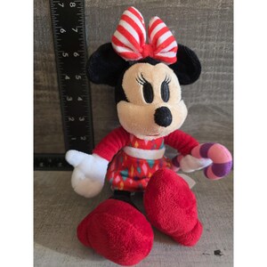 May include: A plush Minnie Mouse doll with a red dress, shoes, and bow. The bow has white and red stripes. The doll is holding a pink and purple candy cane. The doll has black ears and a black nose.