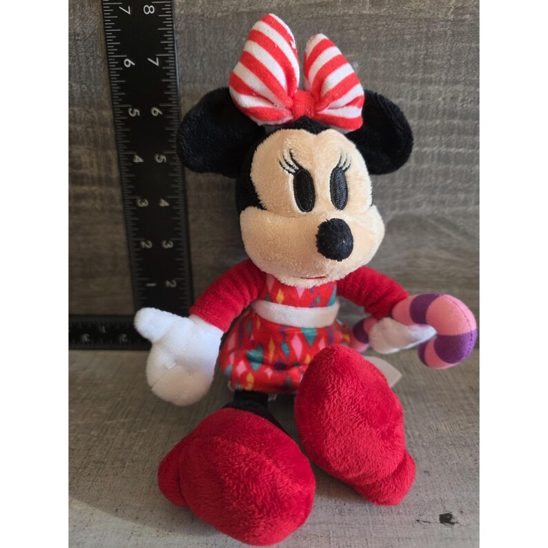 May include: A plush Minnie Mouse doll with a red dress, shoes, and bow. The bow has white and red stripes. The doll is holding a pink and purple candy cane. The doll has black ears and a black nose.
