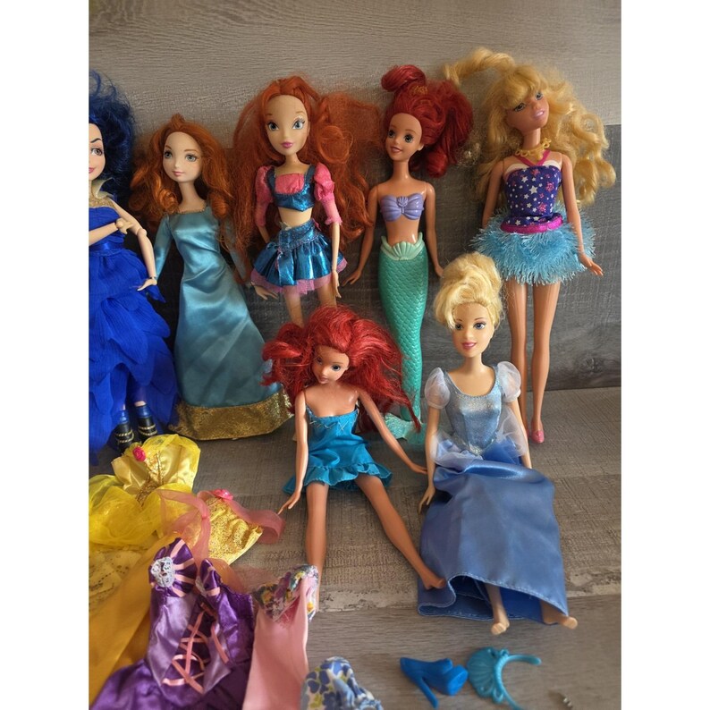 Op de afbeelding: Een verzameling modepoppen met verschillende kapsels en outfits. De poppen dragen een verscheidenheid aan kleurrijke jurken en jurken, waaronder blauw, roze en geel. Sommige poppen hebben zeemeerminstaarten. De poppen zijn gerangschikt op een lichtgekleurd oppervlak.