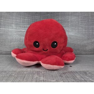 May include: A plush octopus toy featuring a vibrant red exterior and a soft light pink underside. The stuffed animal has two large, round black embroidered eyes and a small, simple black embroidered mouth centered on its rounded head.
