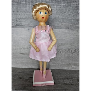 May include: A wooden ballerina doll with blonde hair, a pink dress, and a crown. The doll is standing on a pink base with the year 2015 printed on it.
