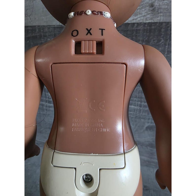 May include: A brown plastic doll with a light tan diaper. The doll has a battery compartment on its back, with the letters "OXT" above a switch. A necklace with white beads is around the doll's neck. The doll is made in China.