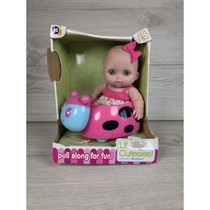 May include: A baby doll with a pink bow in its hair, wearing a floral top, seated inside a pink and blue ladybug-shaped pull-along toy. The packaging features the text Lil' Cutesies! and pull along for fun! with a JC Toys logo at the top.