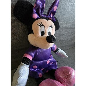 Op de afbeelding: Een pluche Minnie Mouse pop, met een zwarte kop, grote oren en een roze strik met paarse stippen. Ze draagt een paarse jurk met roze stippen en roze rand. Haar handen zijn wit en haar voeten zijn roze.