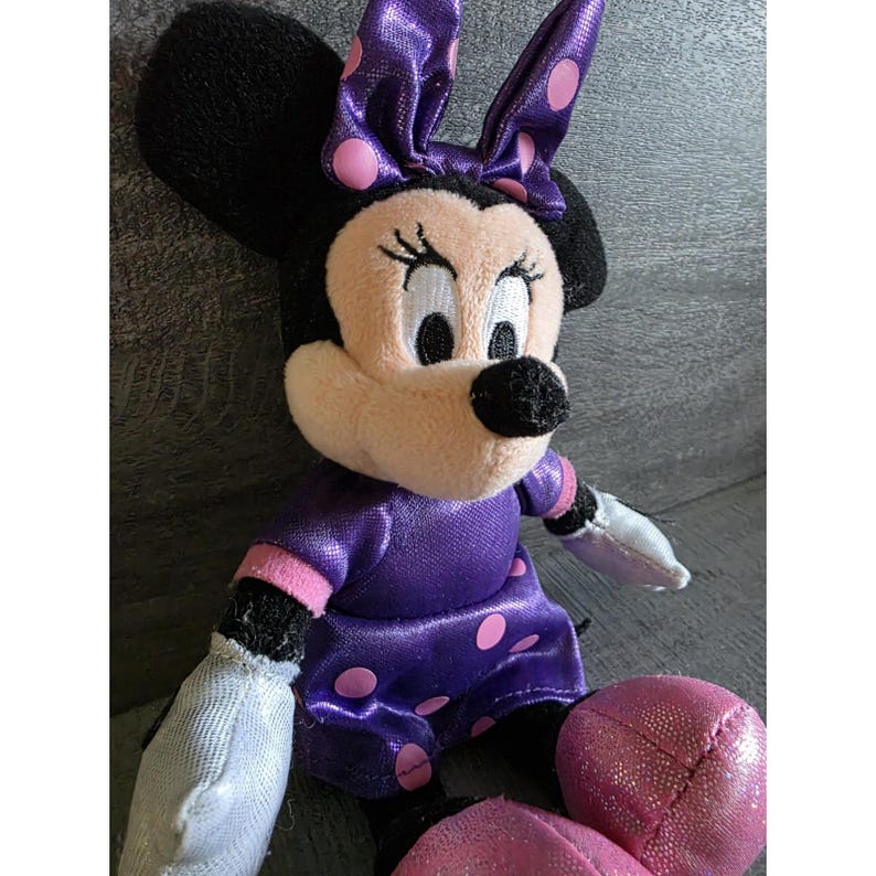 Op de afbeelding: Een pluche Minnie Mouse pop, met een zwarte kop, grote oren en een roze strik met paarse stippen. Ze draagt een paarse jurk met roze stippen en roze rand. Haar handen zijn wit en haar voeten zijn roze.