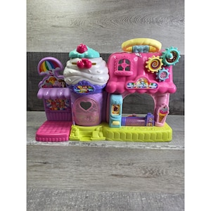 May include: A colorful plastic toy playset shaped like a sweet shop. The playset features a cupcake, ice cream cone, and shop with the text "Sweet Surprise Treat Shoppe". The toy is primarily pink, purple, and yellow.