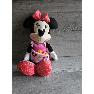 May include: A plush Minnie Mouse toy wearing a pink dress with a yellow sash and a red bow. The toy has black eyes, a black nose, and a pink mouth. The toy is sitting on a wooden surface.