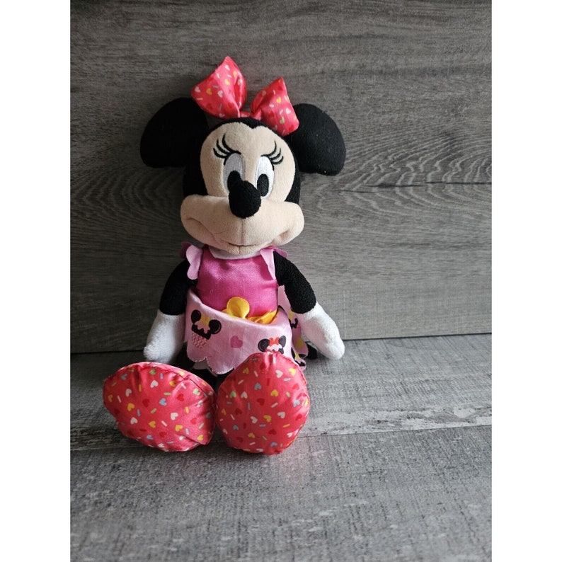 May include: A plush Minnie Mouse toy wearing a pink dress with a yellow sash and a red bow. The toy has black eyes, a black nose, and a pink mouth. The toy is sitting on a wooden surface.