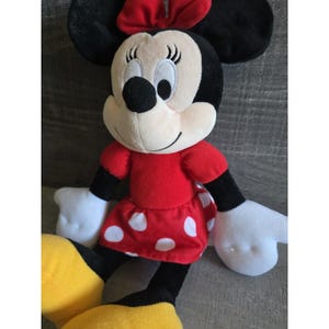 May include: A plush Minnie Mouse doll. The doll has a black head and ears, a white face, and a red bow. She wears a red dress with white polka dots, black arms and legs, and yellow shoes. The doll is soft and cuddly.