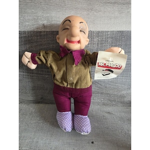 Puede incluir: Muñeco de peluche de Mr. Magoo con cabeza calva, nariz grande y expresión sonriente. El muñeco lleva una camisa marrón, pantalones morados y zapatos de lunares lavanda. Una etiqueta dice "Original Mr. Magoo."
