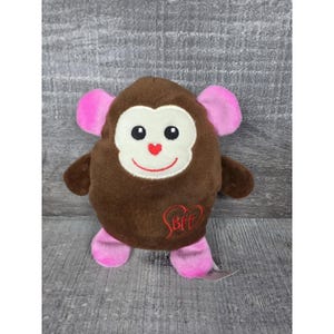 May include: A brown plush monkey toy with pink ears and feet. The monkey has a cream-colored face, black embroidered eyes, a red heart-shaped nose, and a red smile. A red embroidered heart with the letters BFF is on the lower front of the body.