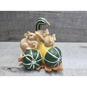 Op de afbeelding: Een grillig beeldje met twee bruine muizen die spelen met een decoratieve kalebas. De kalebas is geel en groen, met een gebogen nek en een gestreept patroon. De muizen bevinden zich bovenop de kalebas en op twee groene pompoenen.
