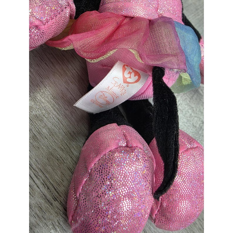 K&ouml;nnte beinhalten: Ein rosa Minnie Mouse Pl&uuml;schtier mit glitzernden Akzenten an F&uuml;&szlig;en und Schleife. Das Spielzeug hat einen rosa T&uuml;llrock und ein Etikett mit der Aufschrift "Sparkle Minnie Disney". Das Pl&uuml;schtier ist schwarz, rosa und wei&szlig;.