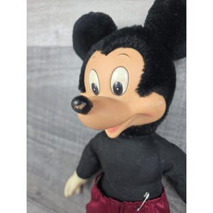 May include: Vintage Mickey Mouse plush doll. The doll features a black body, black ears, and a tan face with large eyes. The doll is wearing maroon pants and a black shirt. A safety pin is attached to the pants.
