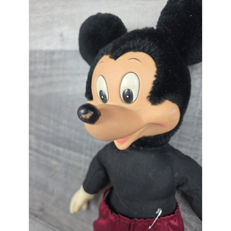 May include: Vintage Mickey Mouse plush doll. The doll features a black body, black ears, and a tan face with large eyes. The doll is wearing maroon pants and a black shirt. A safety pin is attached to the pants.