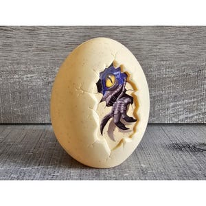 May include: A white ceramic egg with a crack revealing a blue and grey dinosaur hatching from inside.