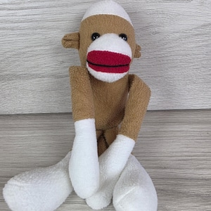 May include: A plush sock monkey toy featuring a brown body with white accents on the head, hands, and feet. The monkey has a prominent white muzzle with a bright red mouth, black stitched eyes, and small brown ears on the sides of its head.
