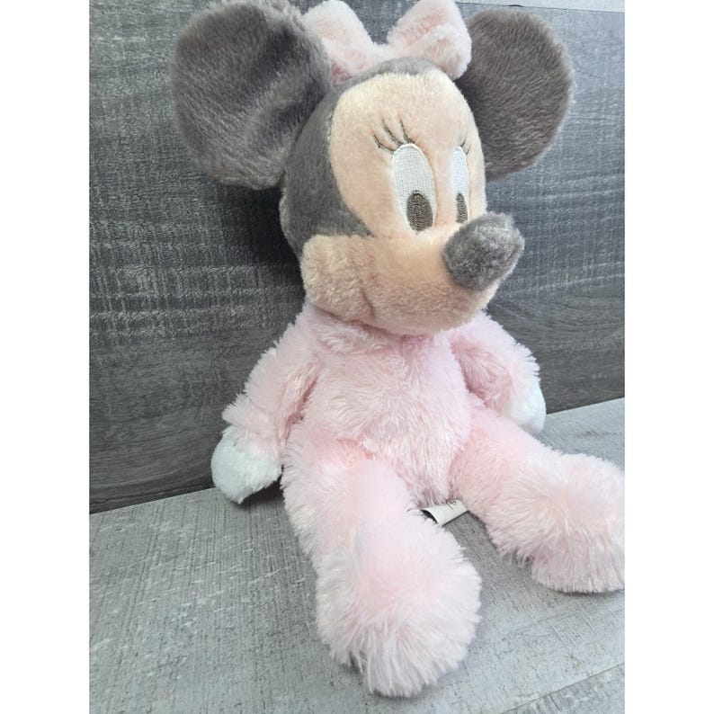 Puede incluir: Un mu&ntilde;eco de peluche de Minnie Mouse con orejas grises y un lazo rosa. El mu&ntilde;eco tiene un cuerpo rosa, detalles blancos en los pies y una nariz gris. El mu&ntilde;eco est&aacute; sentado sobre una superficie gris.