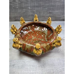 May include: A ceramic bowl with a reddish-brown interior and a speckled green and brown pattern. The rim is adorned with eleven yellow frog figurines. The bowl has a unique, handcrafted appearance.