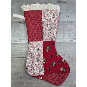 May include: A quilted Christmas stocking with a white lace trim and a white ribbon loop. The stocking features a patchwork design with red and white fabrics, adorned with candy canes, Christmas trees, and teddy bears.