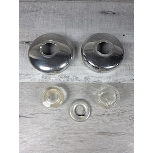 Puede incluir: Dos placas circulares de cromo pulido con aberturas centrales, acompañadas de dos pequeños pomos de cristal transparente y un anillo metálico circular más pequeño, dispuestos sobre una superficie de madera gris claro.
