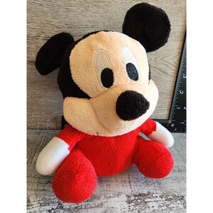 以下が含まれることがあります： 赤と黒のぬいぐるみミッキーマウスの玩具。おもちゃは足を組んで座っており、白い顔に黒い目と黒い鼻があります。