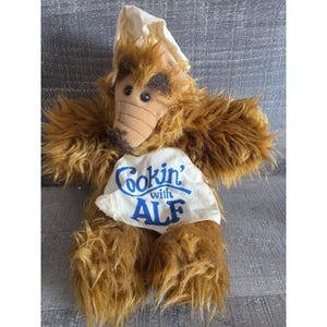 May include: A plush toy of the character Alf, with brown fur and a tan snout. Alf wears a chef's hat and a white apron that reads "Cookin' with ALF" in blue lettering. The toy has large, dark eyes and a friendly expression.