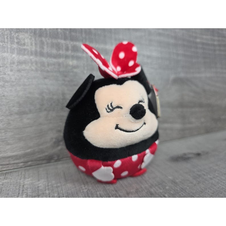 May include: A small, round plush toy featuring a black and cream-coloured character with a winking expression. The toy wears a red dress with white polka dots and a matching red bow with white polka dots on top of its head, complete with black ears.