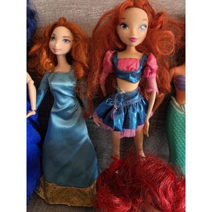 Op de afbeelding: Een verzameling poppen met rood haar en verschillende outfits. E&eacute;n pop draagt een blauwe jurk met gouden rand, terwijl een andere een blauwe en roze top en rok heeft. De poppen staan in verschillende poses.