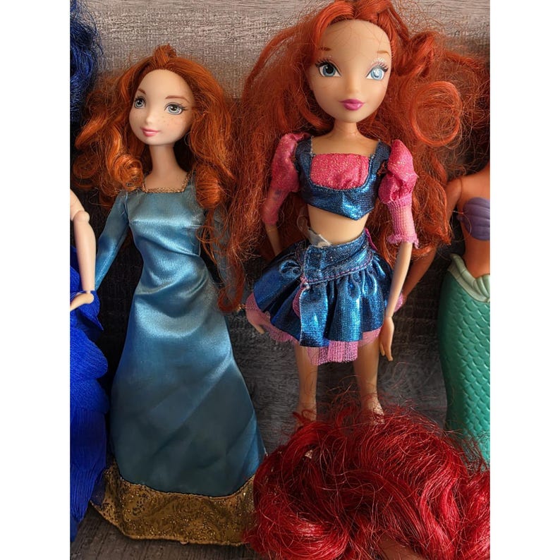 Op de afbeelding: Een verzameling poppen met rood haar en verschillende outfits. E&eacute;n pop draagt een blauwe jurk met gouden rand, terwijl een andere een blauwe en roze top en rok heeft. De poppen staan in verschillende poses.