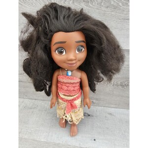 Puede incluir: Una mu&ntilde;eca de piel morena con el pelo largo y negro rizado que lleva un top rojo y blanco y una falda beige con un cintur&oacute;n marr&oacute;n. La mu&ntilde;eca lleva un collar azul con una piedra blanca.