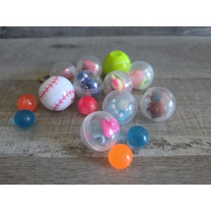 Puede incluir: Una colección de cápsulas de plástico transparente, cada una con un pequeño juguete. Algunas cápsulas están llenas de bolas de colores, mientras que otras contienen figuras en miniatura. También hay una cápsula con temática de béisbol. Las cápsulas están esparcidas sobre una superficie de madera.