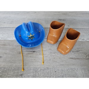 Puede incluir: Un sombrero vaquero de plástico azul con una insignia de estrella de sheriff dorada y una cuerda amarilla. Dos botas vaqueras de plástico marrón.