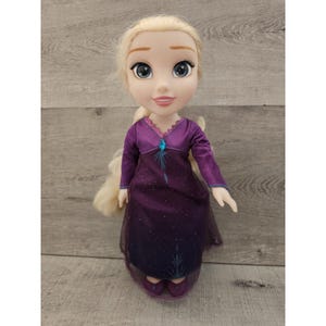 May include: A blonde doll wearing a purple dress with a blue snowflake design. The doll has blue eyes and a smiling face.