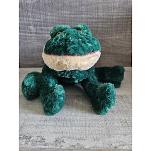 May include: A plush frog toy in shades of green, with a cream-colored mouth and a red line accentuating the lips. The frog has yellow eyes and is made of a soft, textured material. The toy is designed to resemble a frog.