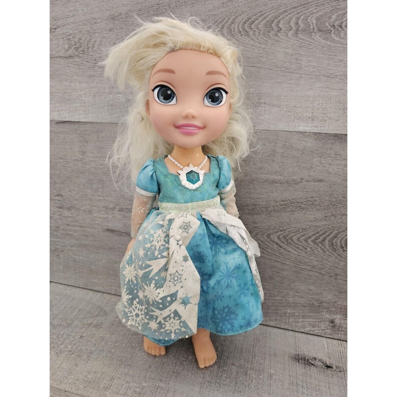 May include: A blonde doll wearing a blue dress with a snowflake pattern. The doll has a light blue necklace with a snowflake pendant. The doll is standing on a wooden surface.