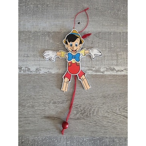 May include: A wooden Pinocchio puppet with a red string and a red bead at the end. The puppet has a yellow shirt, red shorts, a blue bow tie, and a yellow hat with a red feather. The puppet's arms and legs are outstretched.