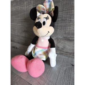 K&ouml;nnte beinhalten: Ein Pl&uuml;schtier von Minnie Mouse, das ein rosa Kleid mit einem bunten Karomuster und einer passenden Schleife tr&auml;gt. Das Spielzeug hat schwarze Ohren und eine schwarze Nase.