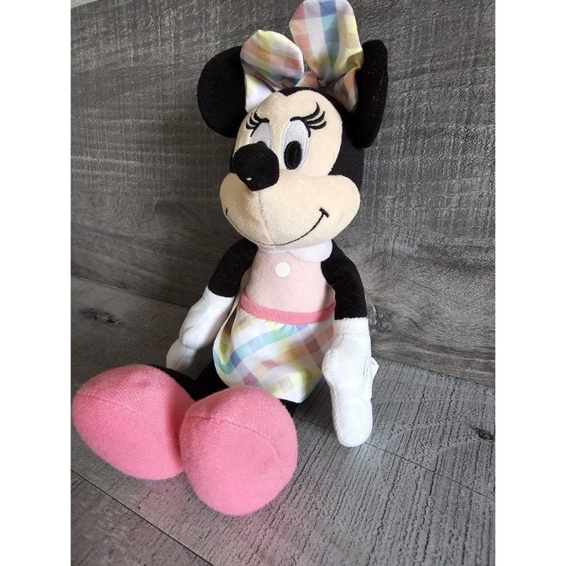 K&ouml;nnte beinhalten: Ein Pl&uuml;schtier von Minnie Mouse, das ein rosa Kleid mit einem bunten Karomuster und einer passenden Schleife tr&auml;gt. Das Spielzeug hat schwarze Ohren und eine schwarze Nase.