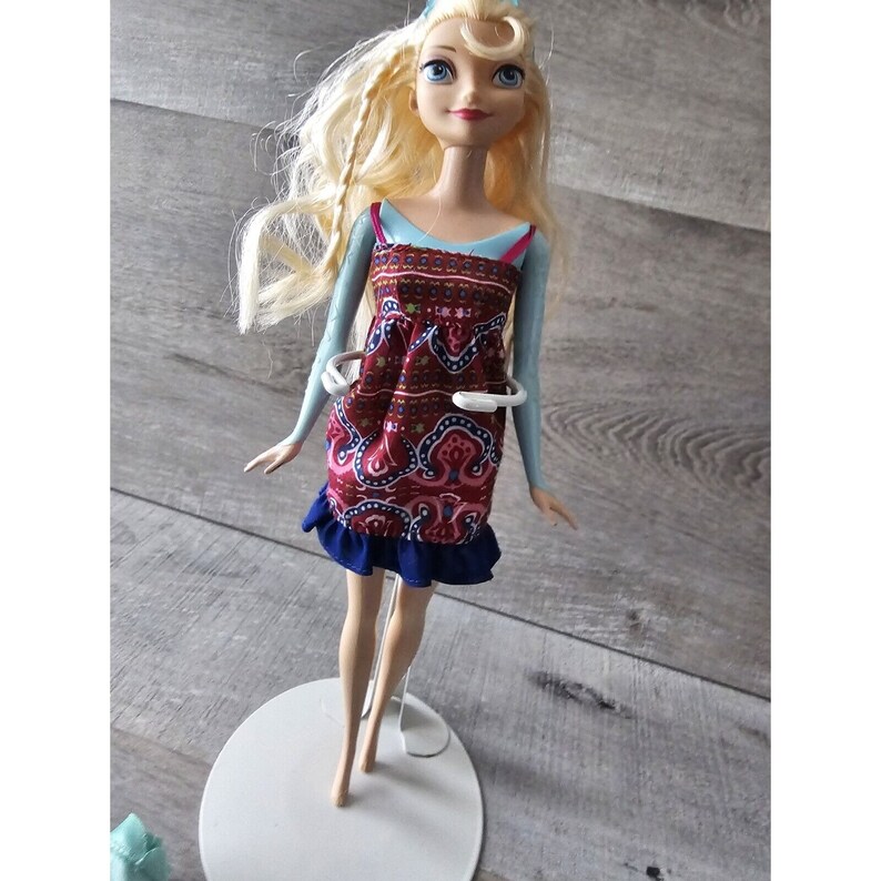 Puede incluir: Una mu&ntilde;eca rubia con un vestido estampado en azul y rojo con un ribete de volantes azules. La mu&ntilde;eca est&aacute; de pie sobre un soporte blanco.