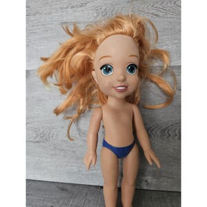 May include: A red-haired doll with blue eyes and freckles wearing a blue swimsuit. The doll has a smiling expression and is posed standing with her arms at her sides.