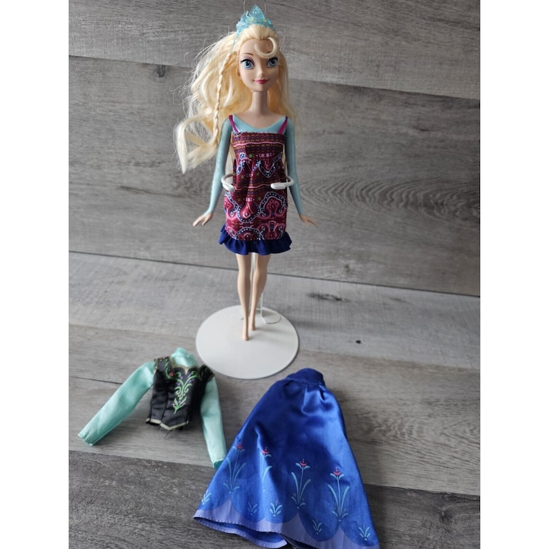 Puede incluir: Una mu&ntilde;eca rubia con un vestido estampado en azul y rojo y una corona. La mu&ntilde;eca est&aacute; de pie sobre un soporte blanco. La mu&ntilde;eca tambi&eacute;n lleva una falda azul y un chaleco verde y negro.