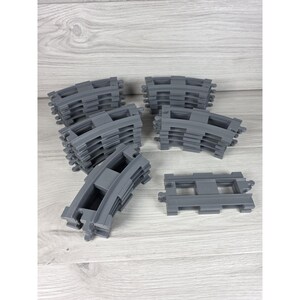 May include: A collection of grey plastic toy train track pieces, including several curved segments stacked in small piles and one straight track piece lying flat, all featuring standard interlocking connectors at each end.