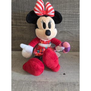 May include: A Minnie Mouse plush toy with a red and white striped bow, black ears, and a red dress with colorful accents. The plush has white gloves, red shoes, and is holding a pink and purple candy cane.