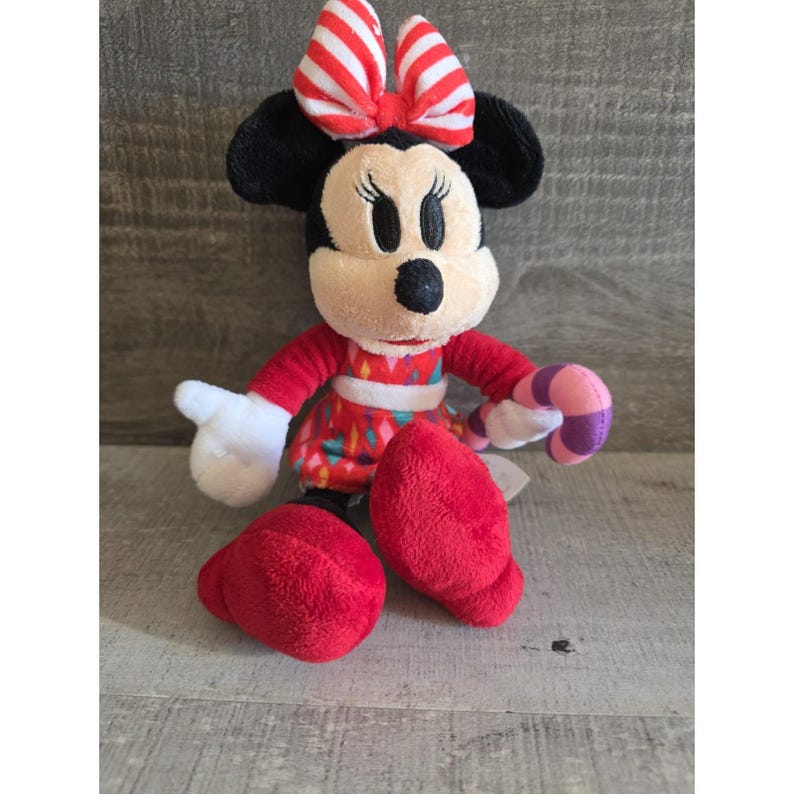 May include: A Minnie Mouse plush toy with a red and white striped bow, black ears, and a red dress with colorful accents. The plush has white gloves, red shoes, and is holding a pink and purple candy cane.