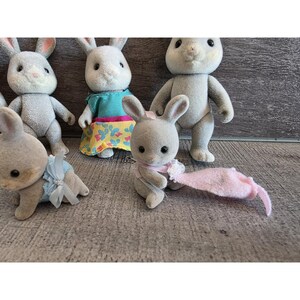 Puede incluir: Una colecci&oacute;n de figuras de conejos grises con ojos negros y orejas forradas de rosa. Algunos conejos visten trajes azules o turquesas, y uno tiene una bufanda rosa y un juguete rosa. Los juguetes est&aacute;n sobre una superficie gris.